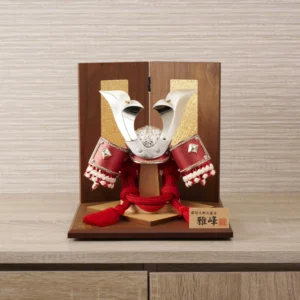 Japanese Kabuto samurai helmet with bold red cords and gold detailing, displayed on a wooden base with folding screen backdrop. Wholesale by Fujiki Crafts.