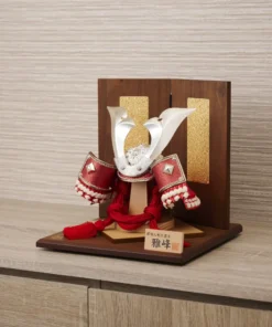 Japanese Kabuto samurai helmet with bold red cords and gold detailing, displayed on a wooden base with folding screen backdrop. Wholesale by Fujiki Crafts.