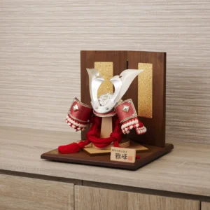 Japanese Kabuto samurai helmet with bold red cords and gold detailing, displayed on a wooden base with folding screen backdrop. Wholesale by Fujiki Crafts.