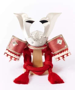 Japanese Kabuto samurai helmet with bold red cords and gold detailing, displayed on a wooden base with folding screen backdrop. Wholesale by Fujiki Crafts.