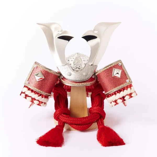 Japanese Kabuto samurai helmet with bold red cords and gold detailing, displayed on a wooden base with folding screen backdrop. Wholesale by Fujiki Crafts.