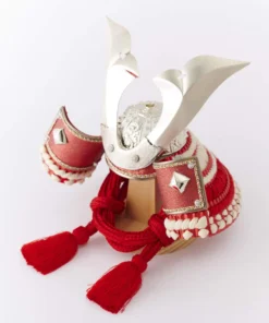 Japanese Kabuto samurai helmet with bold red cords and gold detailing, displayed on a wooden base with folding screen backdrop. Wholesale by Fujiki Crafts.