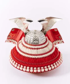 Japanese Kabuto samurai helmet with bold red cords and gold detailing, displayed on a wooden base with folding screen backdrop. Wholesale by Fujiki Crafts.
