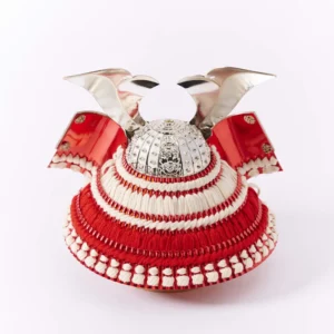 Japanese Kabuto samurai helmet with bold red cords and gold detailing, displayed on a wooden base with folding screen backdrop. Wholesale by Fujiki Crafts.