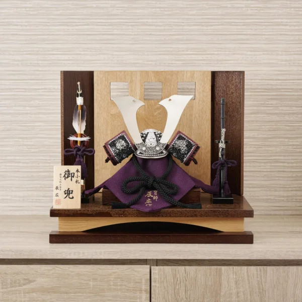 Traditional Kabuto samurai helmet with silver crest, mounted on a dark wood base, flanked by miniature sword and arrow display. Wholesale by Fujiki Crafts.