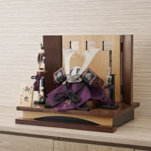 Traditional Kabuto samurai helmet with silver crest, mounted on a dark wood base, flanked by miniature sword and arrow display. Wholesale by Fujiki Crafts.