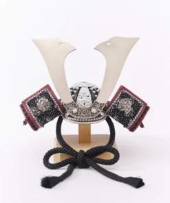 Traditional Kabuto samurai helmet with silver crest, mounted on a dark wood base, flanked by miniature sword and arrow display. Wholesale by Fujiki Crafts.