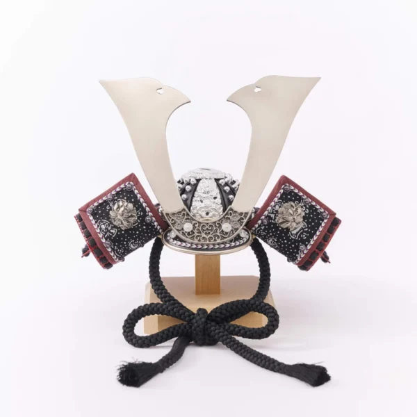 Traditional Kabuto samurai helmet with silver crest, mounted on a dark wood base, flanked by miniature sword and arrow display. Wholesale by Fujiki Crafts.