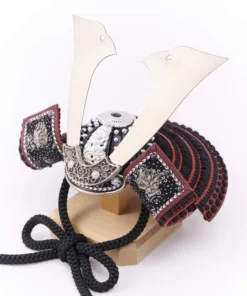 Traditional Kabuto samurai helmet with silver crest, mounted on a dark wood base, flanked by miniature sword and arrow display. Wholesale by Fujiki Crafts.