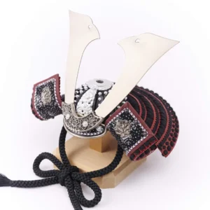 Traditional Kabuto samurai helmet with silver crest, mounted on a dark wood base, flanked by miniature sword and arrow display. Wholesale by Fujiki Crafts.