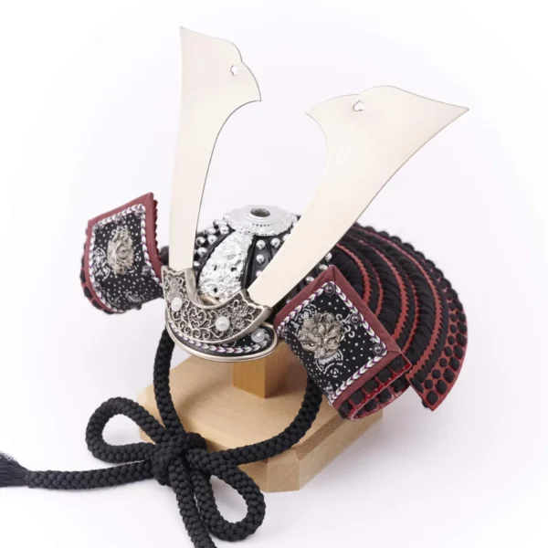 Traditional Kabuto samurai helmet with silver crest, mounted on a dark wood base, flanked by miniature sword and arrow display. Wholesale by Fujiki Crafts.