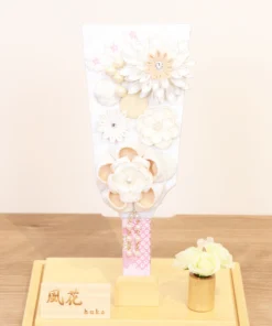 Elegant handcrafted Hagoita paddle adorned with white and beige Kanzashi flowers, set in a wooden base with acrylic case and soft floral accent. Wholesale by Fujikicrafts.