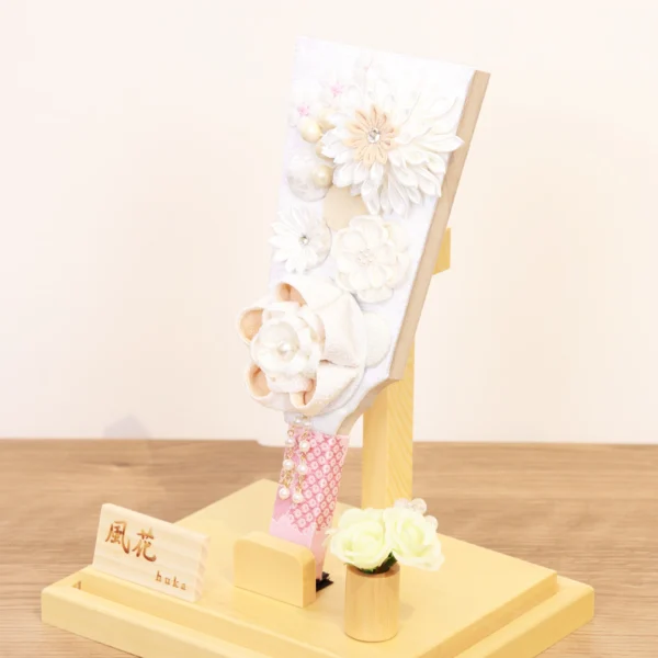 Elegant handcrafted Hagoita paddle adorned with white and beige Kanzashi flowers, set in a wooden base with acrylic case and soft floral accent. Wholesale by Fujikicrafts.