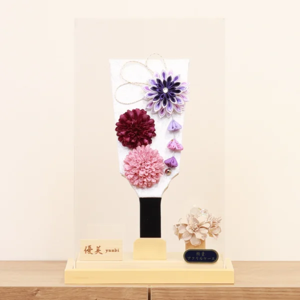 Handcrafted Hagoita paddle with purple, burgundy, and blush pink Kanzashi flowers, accented with trailing petals and displayed in a wooden base with a clear acrylic case. Wholesale by Fujiki Crafts.
