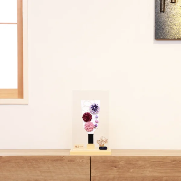 Handcrafted Hagoita paddle with purple, burgundy, and blush pink Kanzashi flowers, accented with trailing petals and displayed in a wooden base with a clear acrylic case. Wholesale by Fujiki Crafts.