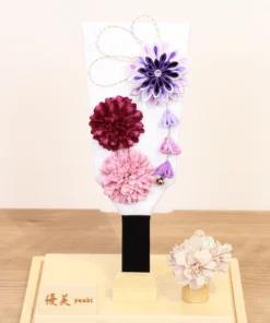 Handcrafted Hagoita paddle with purple, burgundy, and blush pink Kanzashi flowers, accented with trailing petals and displayed in a wooden base with a clear acrylic case. Wholesale by Fujiki Crafts.