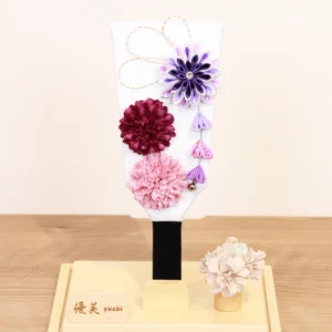 Handcrafted Hagoita paddle with purple, burgundy, and blush pink Kanzashi flowers, accented with trailing petals and displayed in a wooden base with a clear acrylic case. Wholesale by Fujiki Crafts.