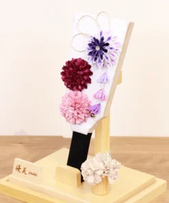 Handcrafted Hagoita paddle with purple, burgundy, and blush pink Kanzashi flowers, accented with trailing petals and displayed in a wooden base with a clear acrylic case. Wholesale by Fujiki Crafts.