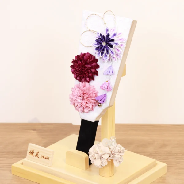Handcrafted Hagoita paddle with purple, burgundy, and blush pink Kanzashi flowers, accented with trailing petals and displayed in a wooden base with a clear acrylic case. Wholesale by Fujiki Crafts.