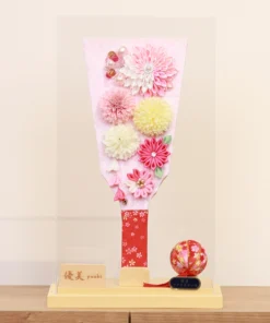 Handmade decorative Hagoita paddle adorned with soft pink, white, and yellow Kanzashi flowers, set in an acrylic case with floral handle and a red temari ball for accent. Wholesale by Fujiki Crafts.