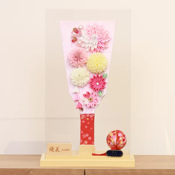 Handmade decorative Hagoita paddle adorned with soft pink, white, and yellow Kanzashi flowers, set in an acrylic case with floral handle and a red temari ball for accent. Wholesale by Fujiki Crafts.