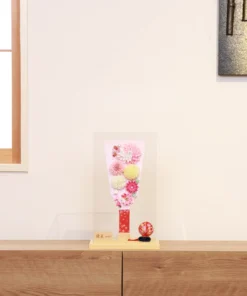 Handmade decorative Hagoita paddle adorned with soft pink, white, and yellow Kanzashi flowers, set in an acrylic case with floral handle and a red temari ball for accent. Wholesale by Fujiki Crafts.