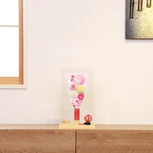 Handmade decorative Hagoita paddle adorned with soft pink, white, and yellow Kanzashi flowers, set in an acrylic case with floral handle and a red temari ball for accent. Wholesale by Fujiki Crafts.