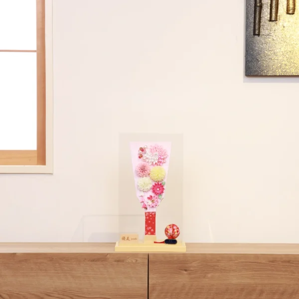 Handmade decorative Hagoita paddle adorned with soft pink, white, and yellow Kanzashi flowers, set in an acrylic case with floral handle and a red temari ball for accent. Wholesale by Fujiki Crafts.