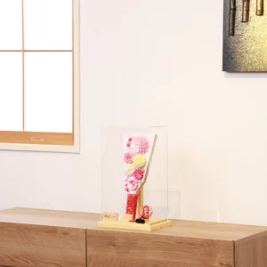 Handmade decorative Hagoita paddle adorned with soft pink, white, and yellow Kanzashi flowers, set in an acrylic case with floral handle and a red temari ball for accent. Wholesale by Fujiki Crafts.