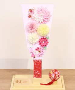 Handmade decorative Hagoita paddle adorned with soft pink, white, and yellow Kanzashi flowers, set in an acrylic case with floral handle and a red temari ball for accent. Wholesale by Fujiki Crafts.