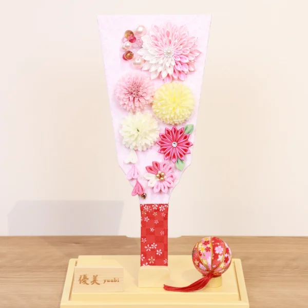 Handmade decorative Hagoita paddle adorned with soft pink, white, and yellow Kanzashi flowers, set in an acrylic case with floral handle and a red temari ball for accent. Wholesale by Fujiki Crafts.