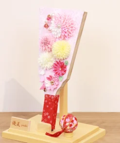 Handmade decorative Hagoita paddle adorned with soft pink, white, and yellow Kanzashi flowers, set in an acrylic case with floral handle and a red temari ball for accent. Wholesale by Fujiki Crafts.
