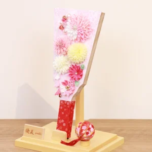 Handmade decorative Hagoita paddle adorned with soft pink, white, and yellow Kanzashi flowers, set in an acrylic case with floral handle and a red temari ball for accent. Wholesale by Fujiki Crafts.