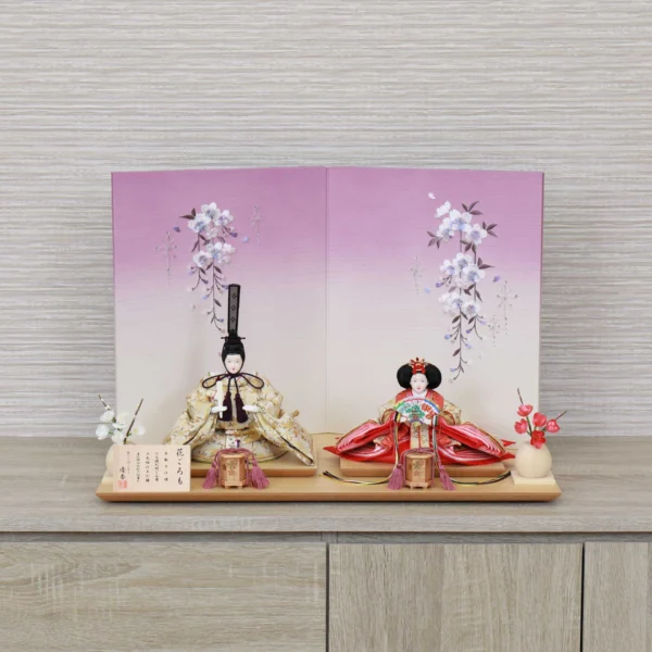 Handcrafted Japanese Hina Doll set with gold and red garments, displayed against a cherry blossom folding screen on wooden platform. Wholesale by Fujiki Crafts.