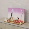Handcrafted Japanese Hina Doll set with gold and red garments, displayed against a cherry blossom folding screen on wooden platform. Wholesale by Fujiki Crafts.