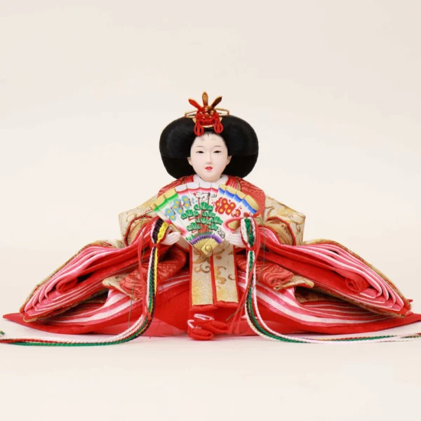 Empress Hina Doll wearing a vivid red kimono with gold detailing, holding a colorful decorative folding fan, and styled with a traditional court hairstyle topped with a red crown. Wholesale by Fujiki Crafts.