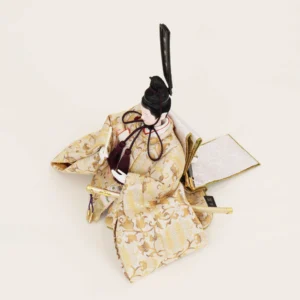 Emperor Hina Doll dressed in a pale gold brocade kimono with intricate patterns, holding a slender fan and wearing a tall black traditional court headdress. Wholesale by Fujiki Crafts.