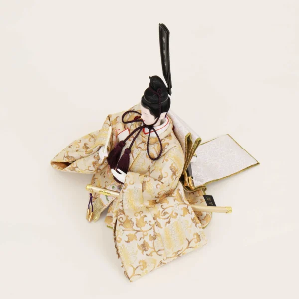 Emperor Hina Doll dressed in a pale gold brocade kimono with intricate patterns, holding a slender fan and wearing a tall black traditional court headdress. Wholesale by Fujiki Crafts.