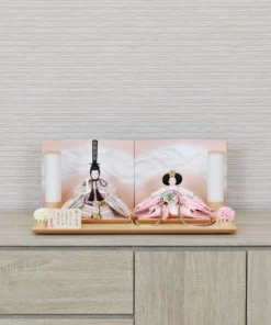 Elegant Hina doll set featuring pastel-striped Emperor and soft pink Empress dolls seated before a gradient peach folding screen, flanked by cylindrical lanterns and floral accents on a wooden display base. Wholesale by Fujiki Crafts.