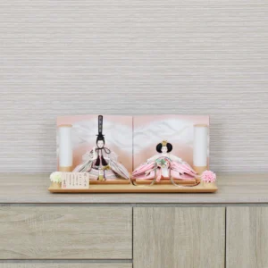 Elegant Hina doll set featuring pastel-striped Emperor and soft pink Empress dolls seated before a gradient peach folding screen, flanked by cylindrical lanterns and floral accents on a wooden display base. Wholesale by Fujiki Crafts.