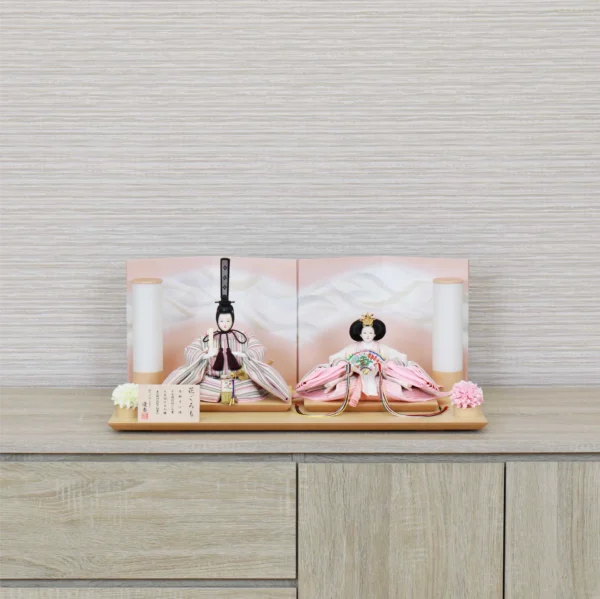 Elegant Hina doll set featuring pastel-striped Emperor and soft pink Empress dolls seated before a gradient peach folding screen, flanked by cylindrical lanterns and floral accents on a wooden display base. Wholesale by Fujiki Crafts.