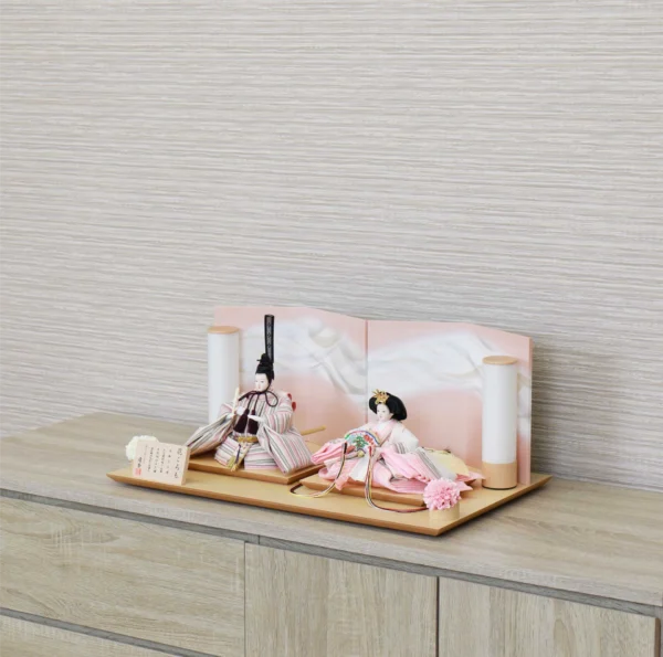 Elegant Hina doll set featuring pastel-striped Emperor and soft pink Empress dolls seated before a gradient peach folding screen, flanked by cylindrical lanterns and floral accents on a wooden display base. Wholesale by Fujiki Crafts.