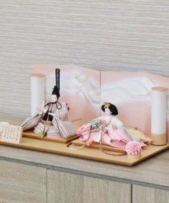 Elegant Hina doll set featuring pastel-striped Emperor and soft pink Empress dolls seated before a gradient peach folding screen, flanked by cylindrical lanterns and floral accents on a wooden display base. Wholesale by Fujiki Crafts.