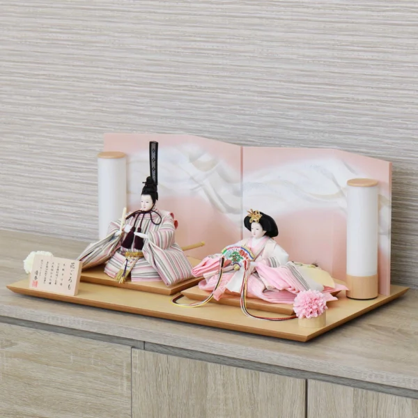 Elegant Hina doll set featuring pastel-striped Emperor and soft pink Empress dolls seated before a gradient peach folding screen, flanked by cylindrical lanterns and floral accents on a wooden display base. Wholesale by Fujiki Crafts.